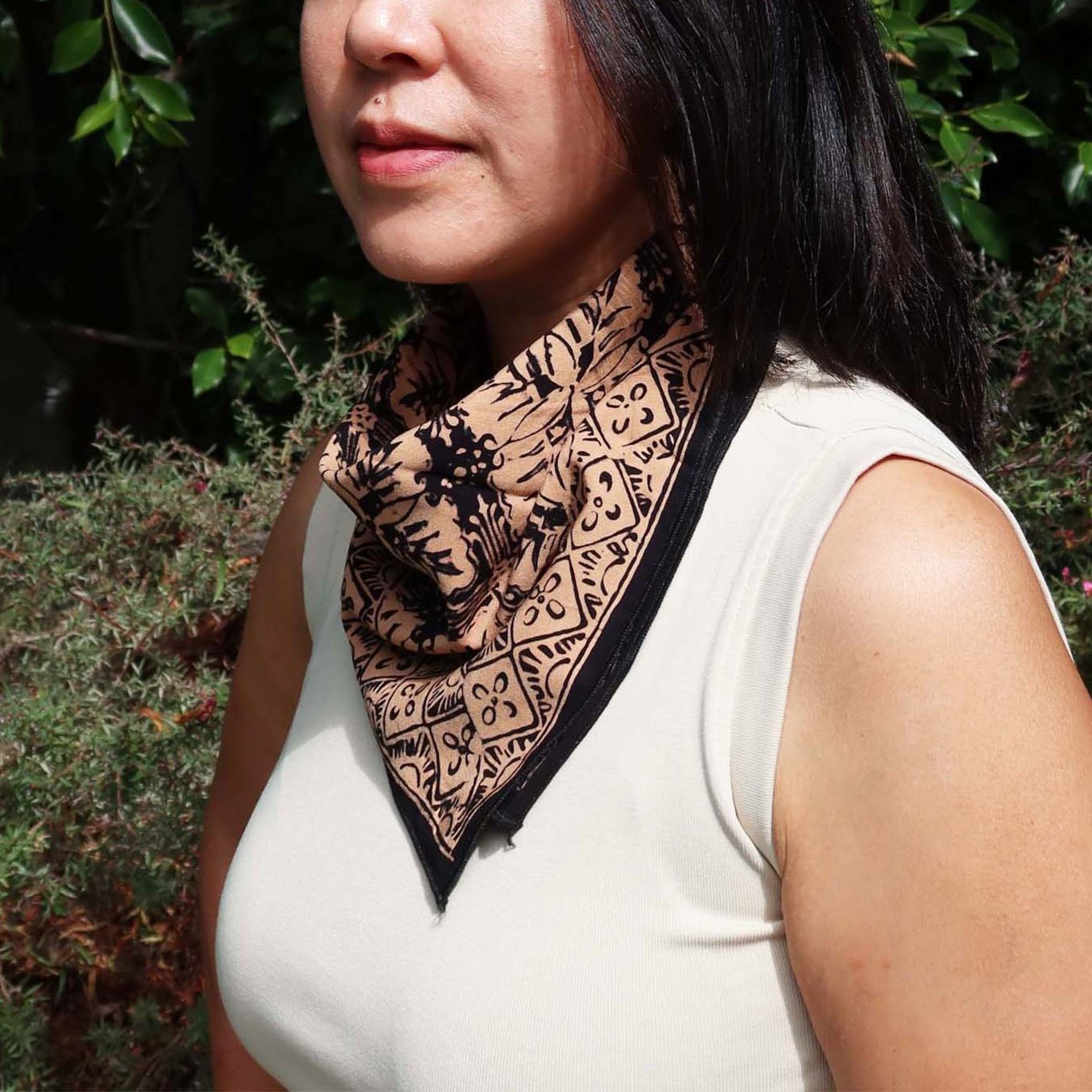 A person wearing a white tank top and a hand-dyed batik kasih co-op cotton bandana in harvest brown and black around their neck.