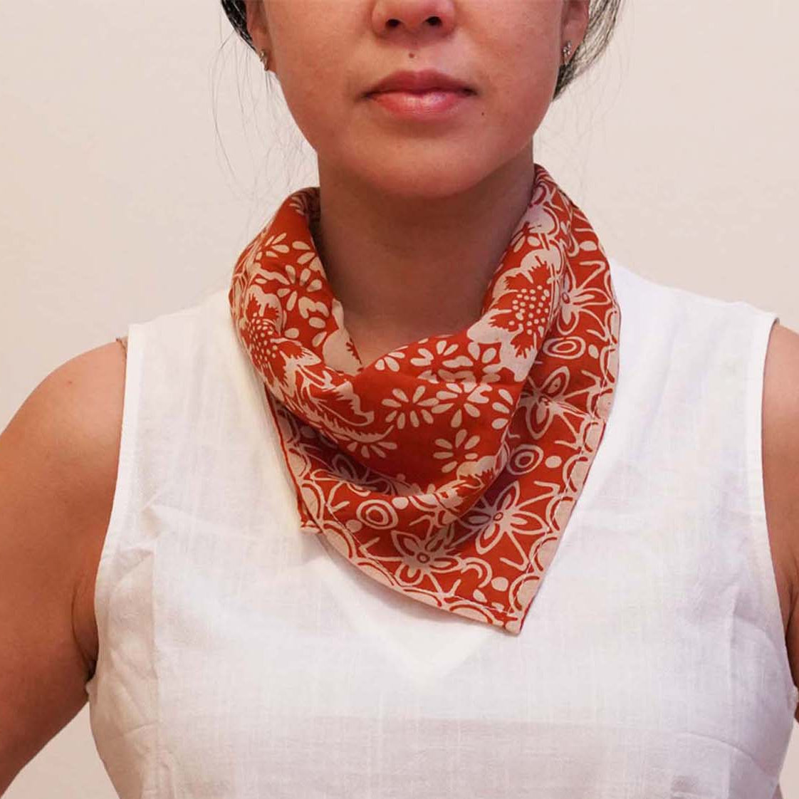 A person wearing a white sleeveless top and white pants, with a red and brown geometric patterned bandana around their neck.