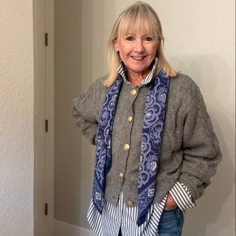 An older woman with blonde hair stands in a hallway wearing a gray knit cardigan over a striped button-up shirt, blue jeans, and a blue paisley patterned scarf.