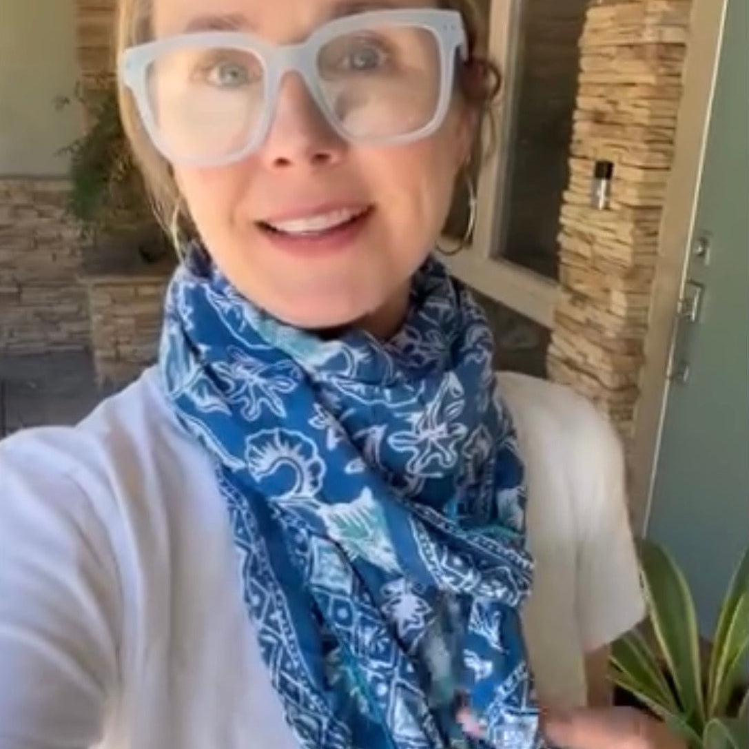 A woman wearing a blue and white paisley bandana scarf and white-framed glasses, taking a selfie in front of a stone pillar and house exterior.