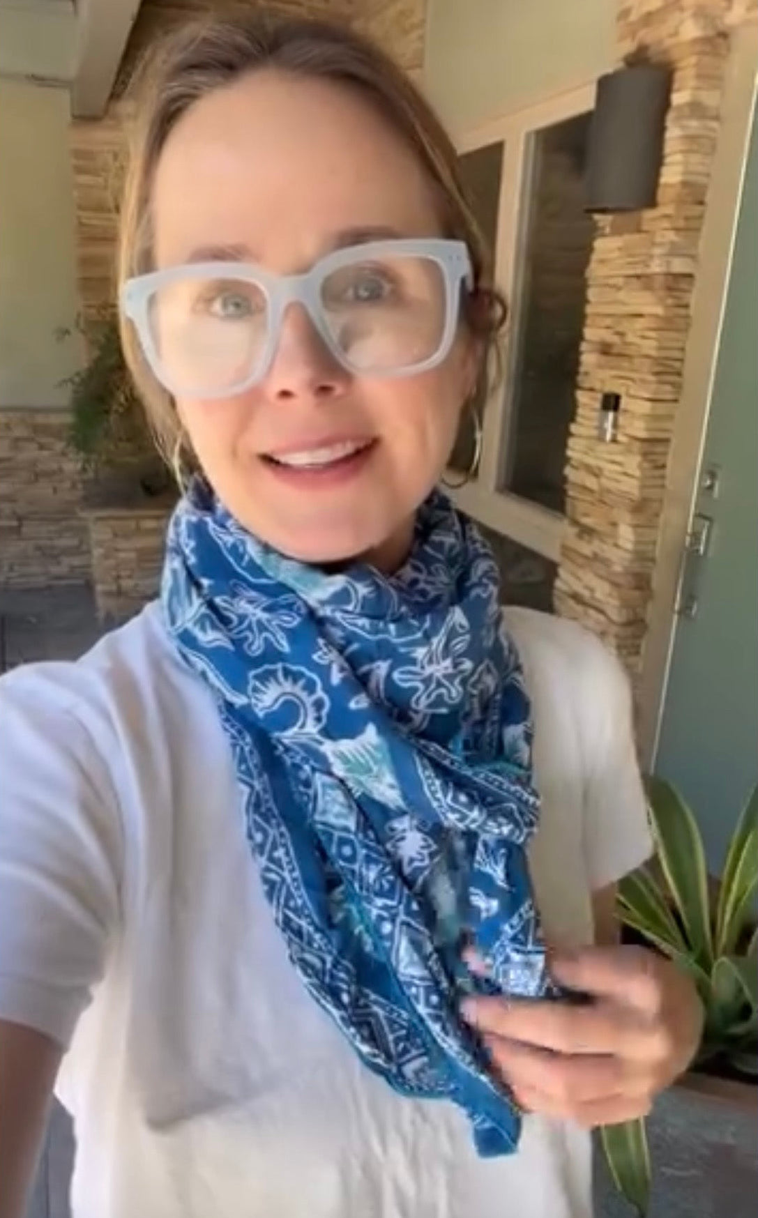 A woman wearing a blue and white paisley bandana scarf and white-framed glasses, taking a selfie in front of a stone pillar and house exterior.