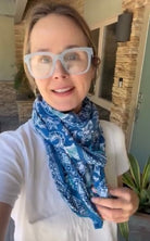A woman wearing a blue and white paisley bandana scarf and white-framed glasses, taking a selfie in front of a stone pillar and house exterior.
